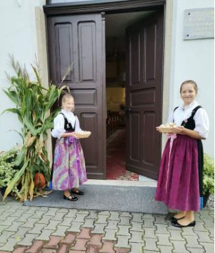 Kaksi hymyilevää tyttöä pitää leipäkoreja käsissään kirkon avoimella ovella. Tytöillä paikalliset juhla-asut.