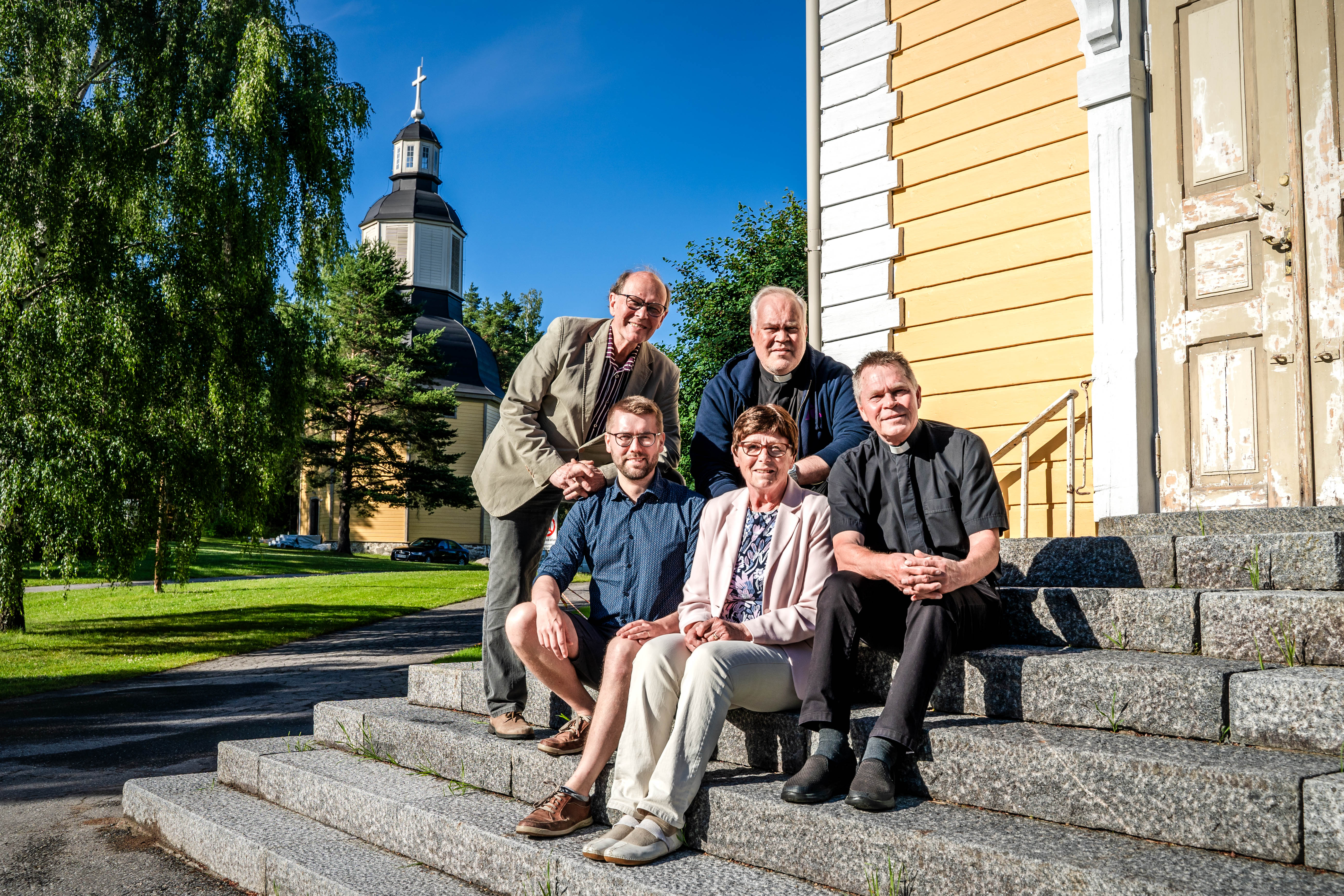 Viisi ihmistä istuu kirkon kiriportailla. Taustalla keltainen puukirkko ja kellotorni.
