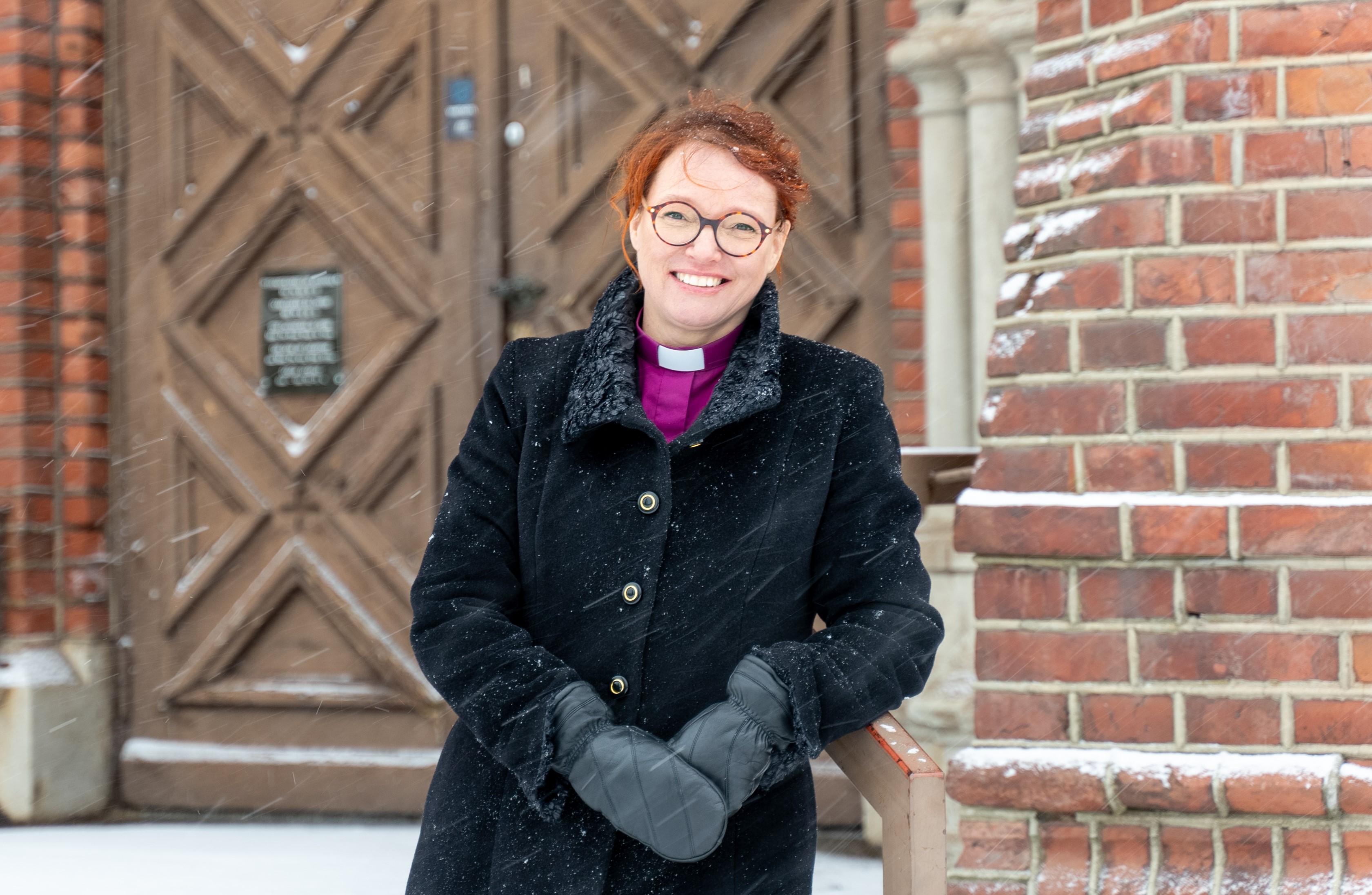 Puolilähikuva naisesta, joka seisoo kirkon edessä lumisateessa.