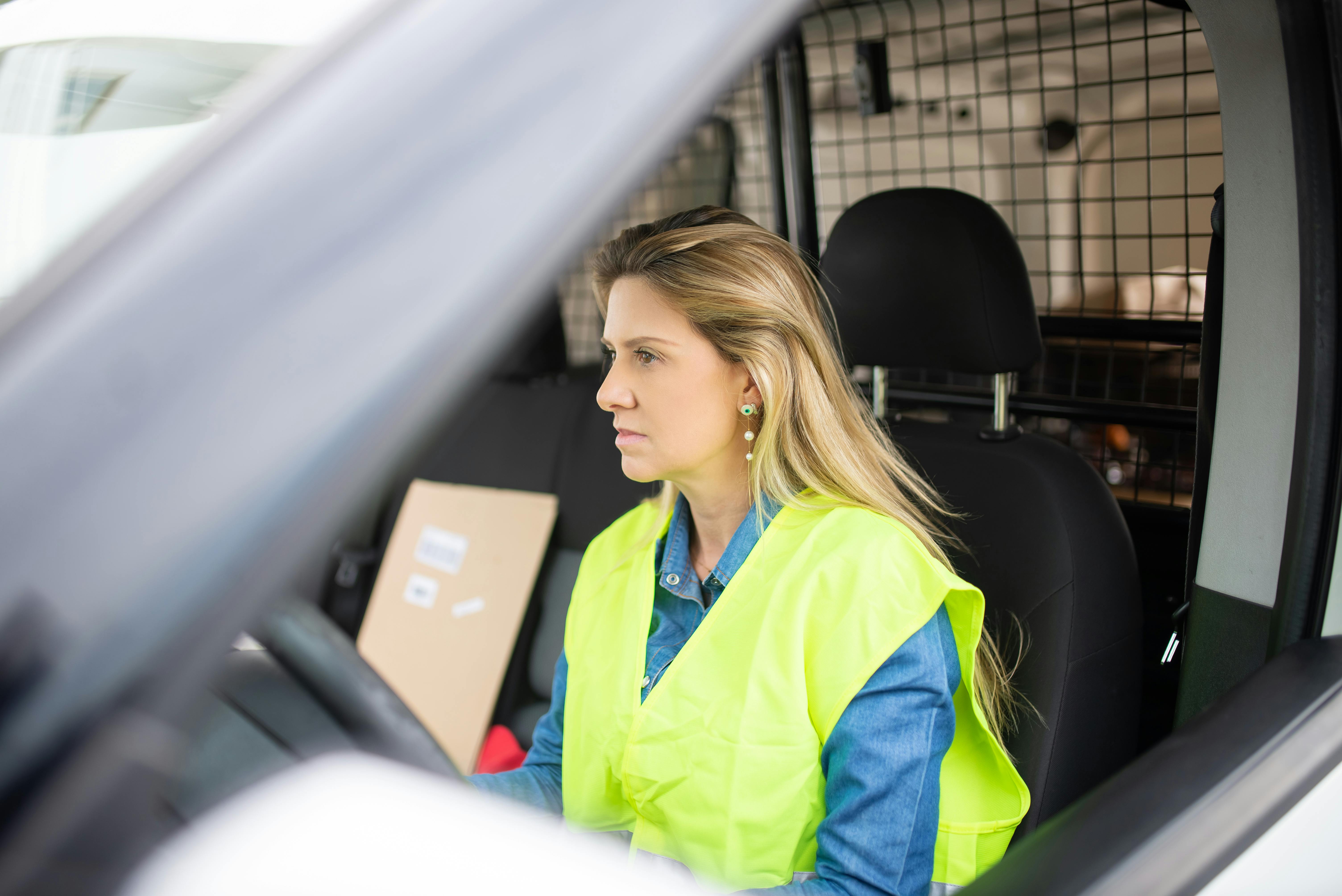 Nainen heijastinliivissä auton ratin takana. Penkillä kirjeitä ja paketteja.
