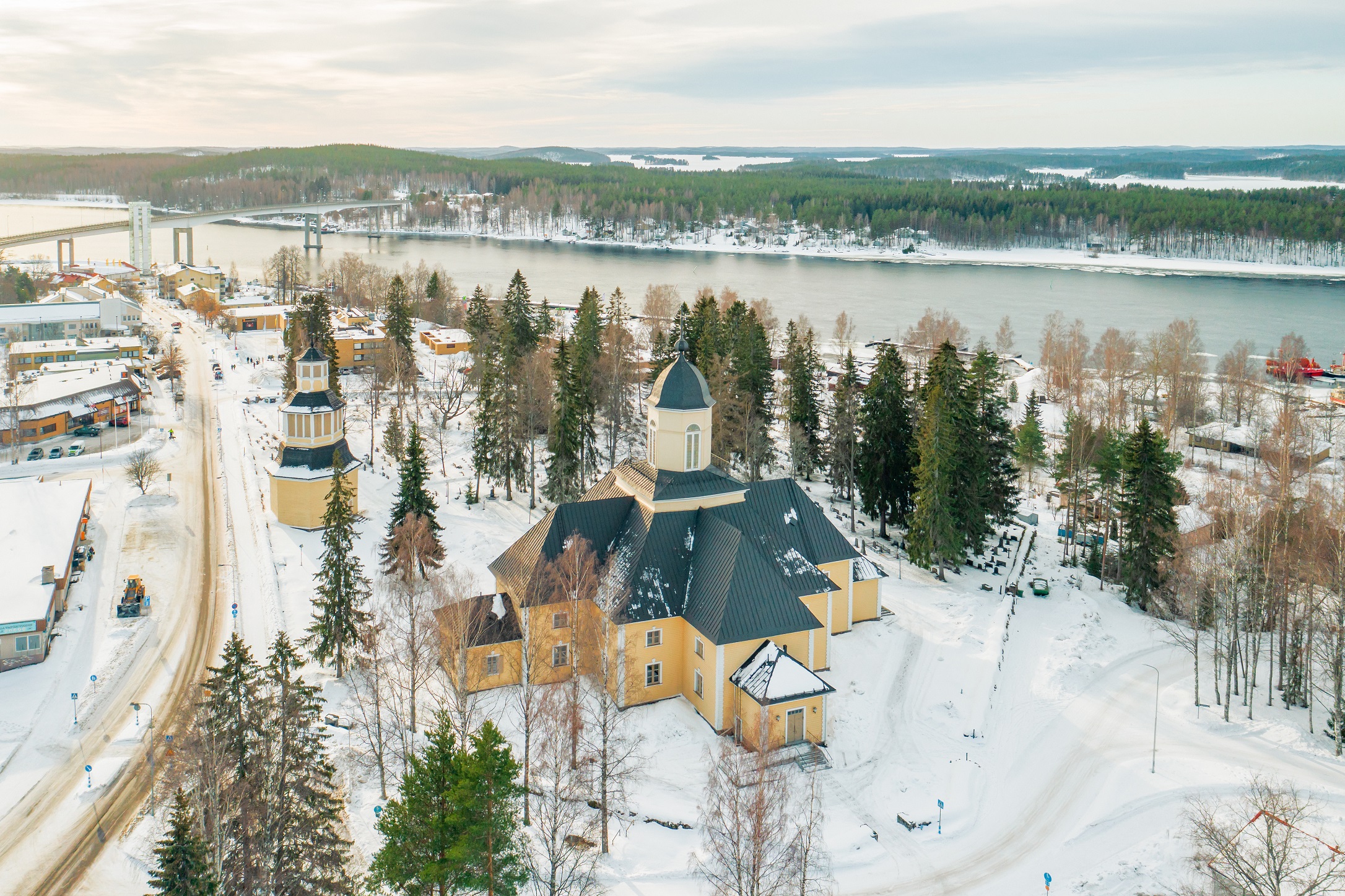 Ilmakuva Puumalan kirkosta ja kellotapulista. Lunta maassa. Taustalla puita ja järvi.