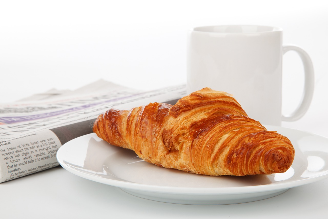 Pöydällä sanomalehti, croissant ja kahvikuppi