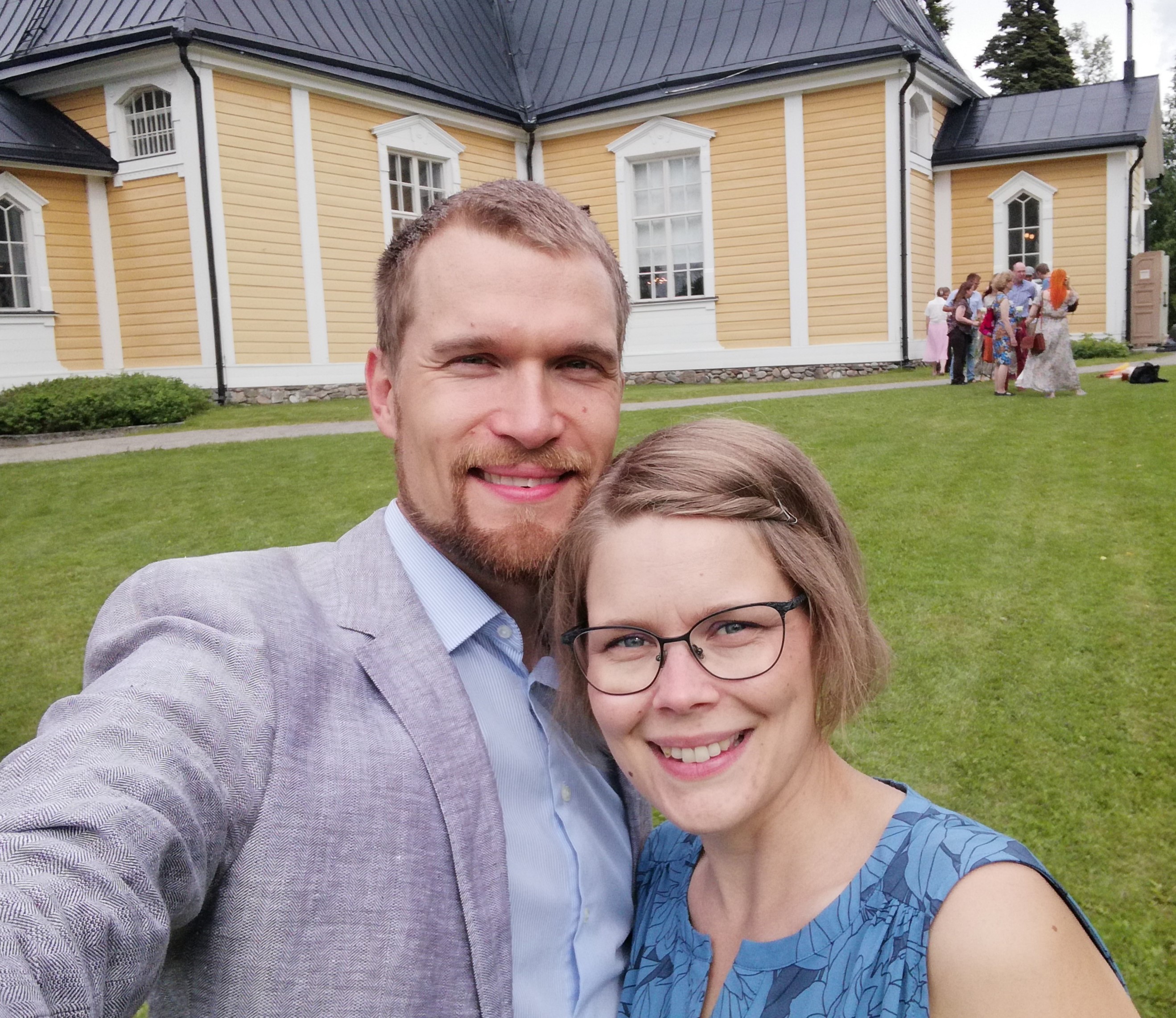 Hymyilevä nuori pariskunta juhlavaatteissaan keltaisen puukirkon edessä.