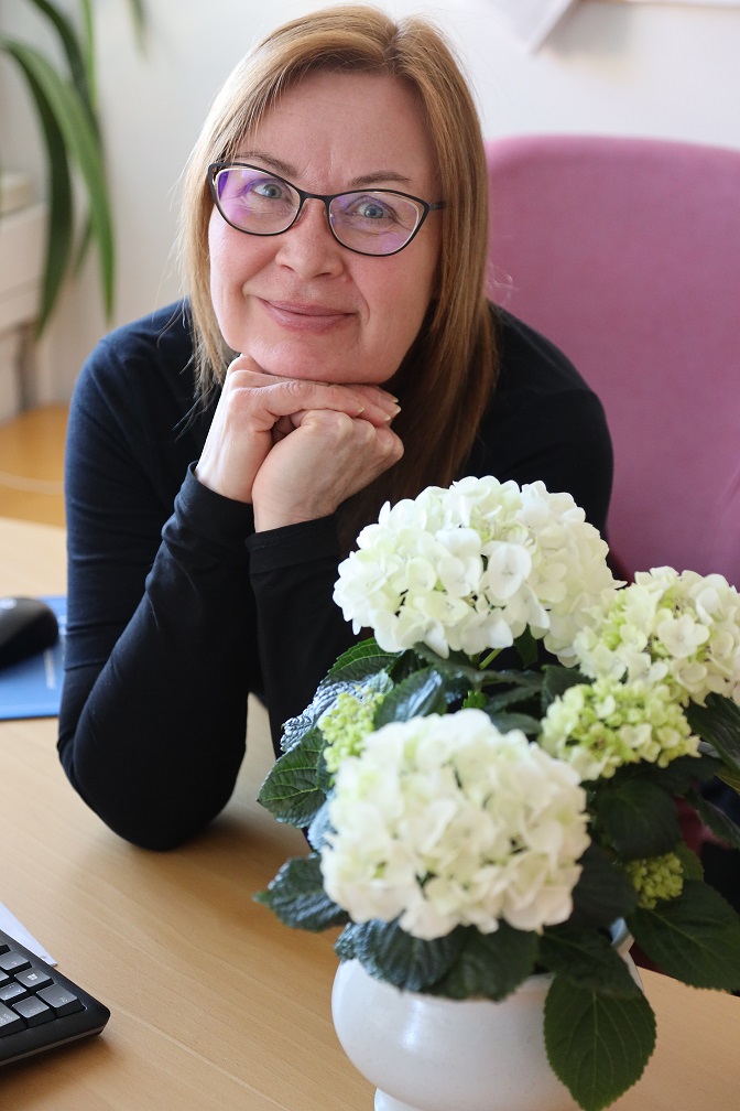Pappi Aija Auren hymyilee katsoen kameraa, etualalla on ihana valkoinen iso kukka.